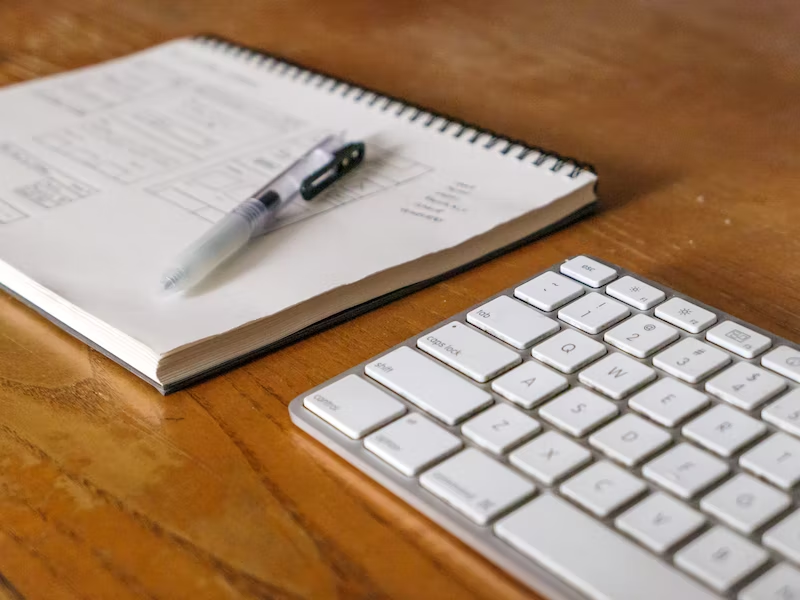 A close up of a keyboard next to a notebook and a pen.
